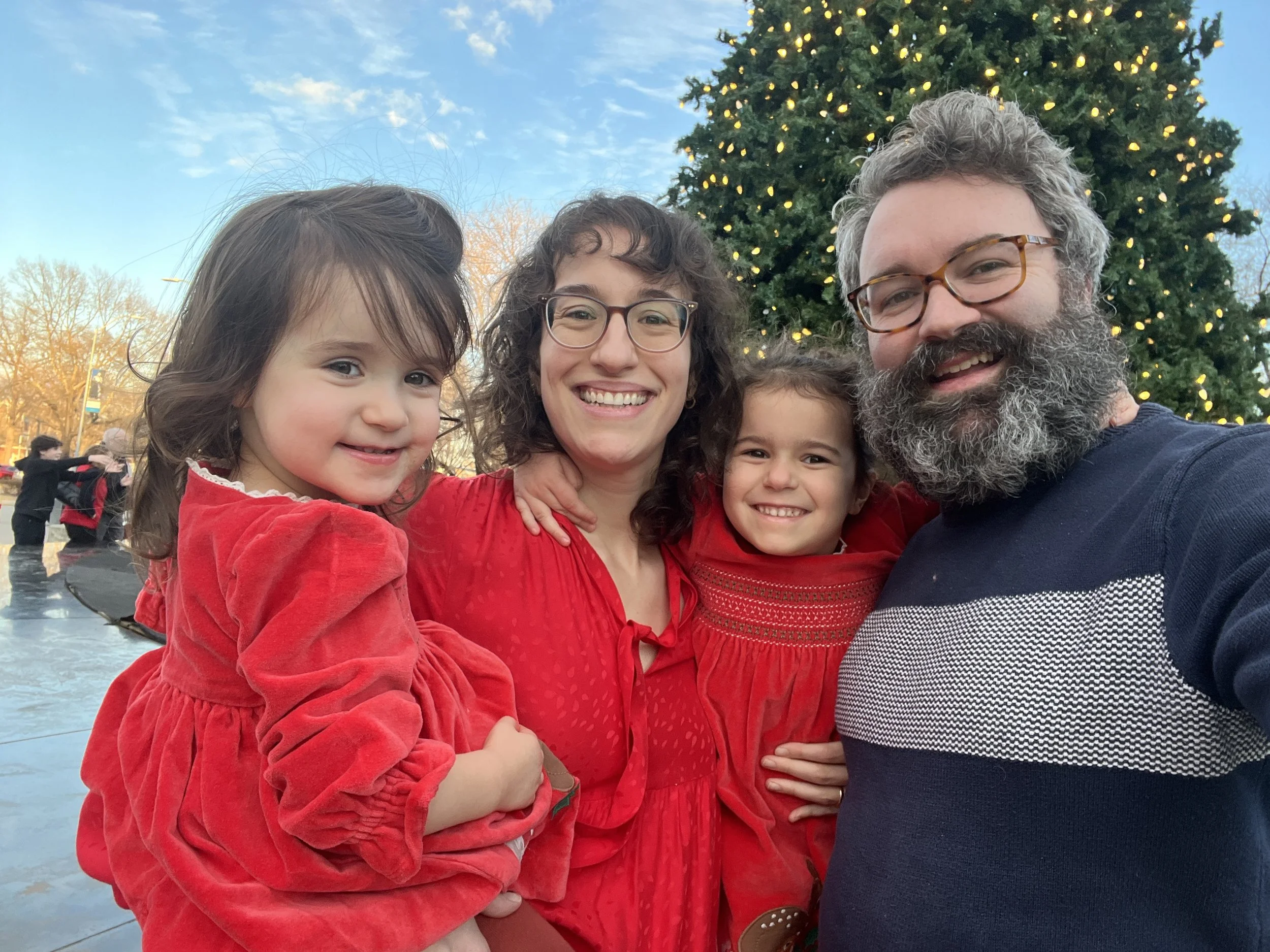 Tom with his wife Lindsey and two of their daughters, all in red, in front of a lit Christmas tree