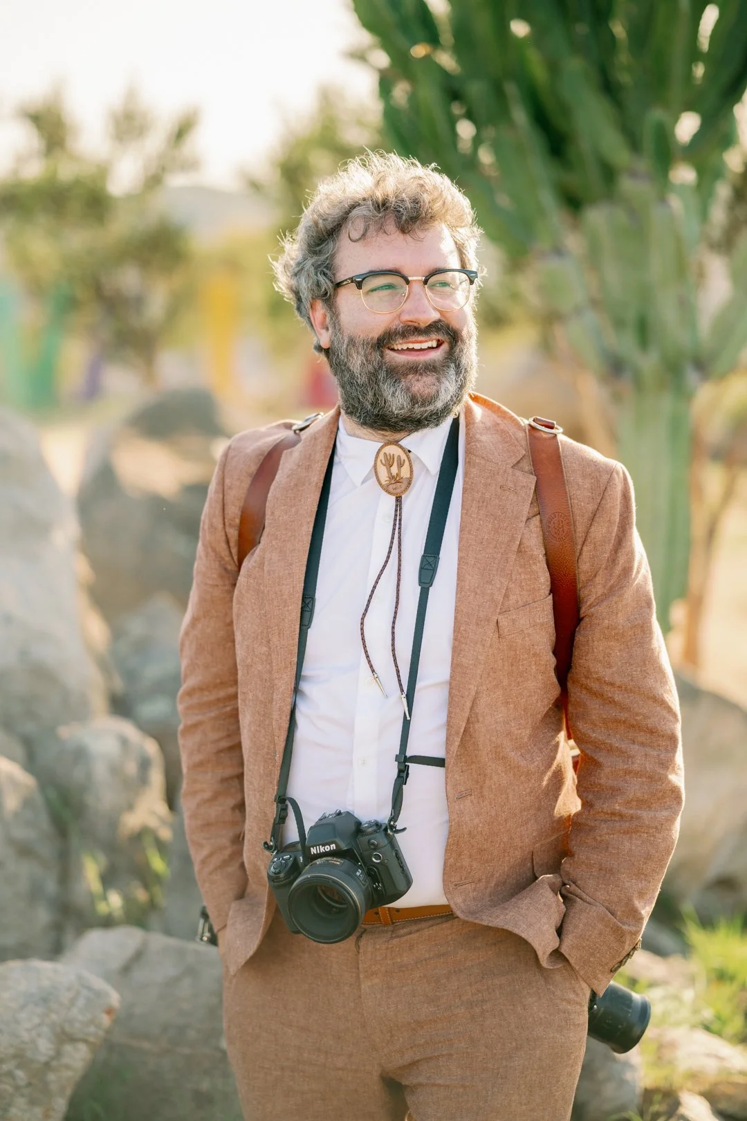 Tom Waldenberg in a rust linen suit and bolo tie, a Nikon camera hanging at his chest